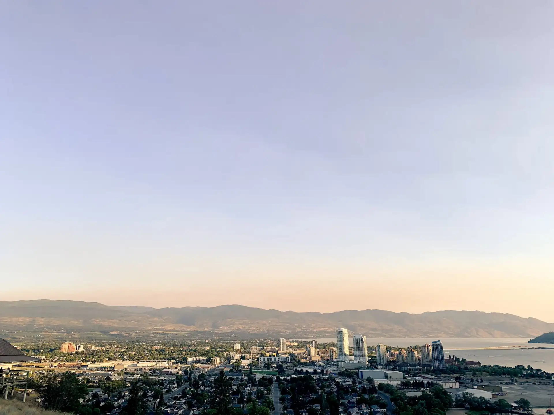 Panoramic view of downtown Kelowna skyline along the shore of Okanagan Lake with surrounding hills in the background.
